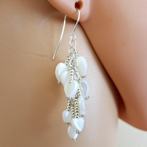 May include: A dangling earring with a silver hook and a cluster of white heart-shaped beads. The beads are strung together with silver-colored metal and small silver beads, creating a cascading effect. The earring is shown against a blurred background.