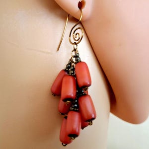 May include: Gold-tone dangle earrings featuring a cluster of coral-colored, oblong beads. The earrings have a spiral design at the top and small black beads between the coral beads. The earrings are shown on a person's ear.