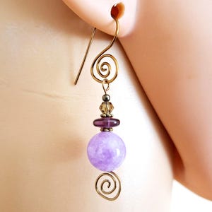May include: A single gold-toned earring with a spiral design and a lavender bead. The earring features a gold hook, a small cluster of beads, and a large, round, light purple bead.