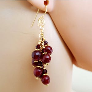 May include: Gold-tone dangle earrings featuring a cluster of deep red, faceted beads. The earrings have a delicate, spiral design at the top and small gold beads accenting the cluster. The earrings are shown against a neutral background.