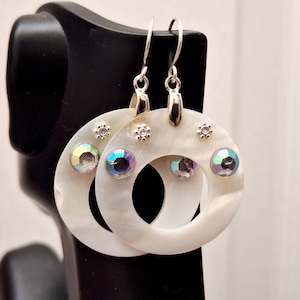May include: A pair of circular drop earrings crafted from white mother-of-pearl. Each earring features a central open circle design accented with three small, iridescent, multifaceted round crystals and three tiny silver-toned metal flower charms.