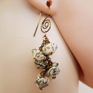 May include: Close-up of a dangle earring with a gold-colored hook and a spiral design. The earring features a cluster of off-white, speckled beads with brown accents and small amber-colored beads.
