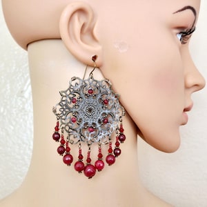 May include: A large, ornate earring with a filigree design in silver and bronze tones. The earring features a central circular design and is adorned with dangling red beads. The earring is designed to be worn in a pierced ear.