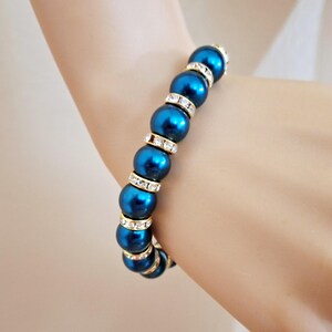 May include: A stretch bracelet featuring alternating round, deep teal blue faux pearls and small, gold-toned spacers encrusted with clear, sparkling rhinestones. The beads are arranged in a repeating pattern, creating a continuous circular design.