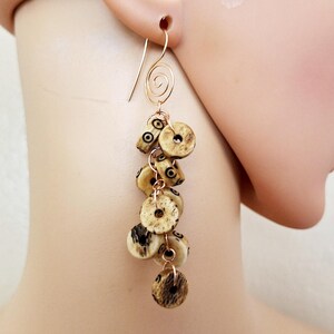 May include: Long dangle earrings featuring a cluster of beige and brown beads with black accents. The earrings are suspended from a gold-colored wire with a spiral design at the top. The beads have a circular shape.