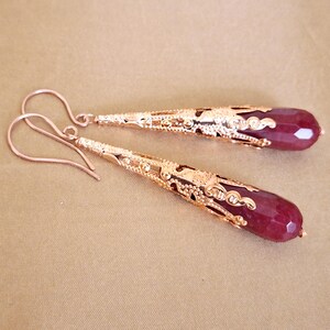 May include: A pair of dangle earrings featuring a teardrop-shaped, faceted, deep red gemstone. The earrings have a gold-colored filigree design that encases the gemstone and a curved ear wire. The earrings are displayed on a neutral background.