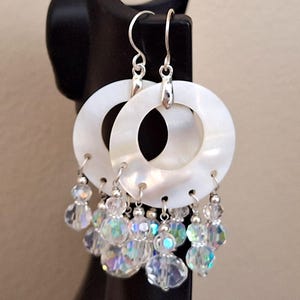 May include: White mother-of-pearl hoop earrings with dangling iridescent crystal beads. The earrings have silver-colored hooks and accents. The circular hoops have a polished, reflective surface. The crystal beads add a touch of sparkle and movement.