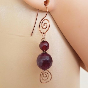 May include: Copper-colored dangle earrings with a spiral design and two dark red, round beads. The earrings are suspended from a copper hook and are shown against a blurred background.