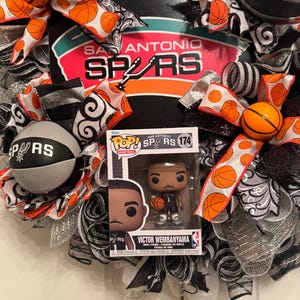 Puede incluir: Una corona decorativa con el logotipo y los colores de los Spurs de San Antonio, con detalles de baloncesto. Incluye una figura Funko Pop! de Victor Wembanyama. La corona est&aacute; adornada con cintas y balones de baloncesto naranjas, negras, blancas y plateadas.