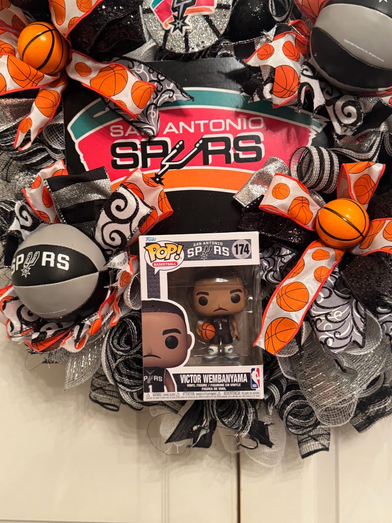 Puede incluir: Una corona decorativa con el logotipo y los colores de los Spurs de San Antonio, con detalles de baloncesto. Incluye una figura Funko Pop! de Victor Wembanyama. La corona est&aacute; adornada con cintas y balones de baloncesto naranjas, negras, blancas y plateadas.