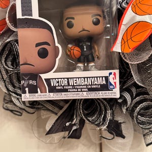 Puede incluir: Una figura de vinilo Funko Pop! de Victor Wembanyama con un uniforme negro y gris de los Spurs, sosteniendo un bal&oacute;n de baloncesto. La figura est&aacute; en una vitrina de pl&aacute;stico transparente. El embalaje incluye el logotipo de la NBA y las palabras "VICTOR WEMBANYAMA".
