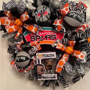 Puede incluir: Una corona decorativa con elementos del equipo de baloncesto de los San Antonio Spurs. La corona incluye cintas con tem&aacute;tica de baloncesto, adornos de baloncesto y una figura Funko Pop de Victor Wembanyama. La combinaci&oacute;n de colores incluye negro, plata, naranja y blanco.