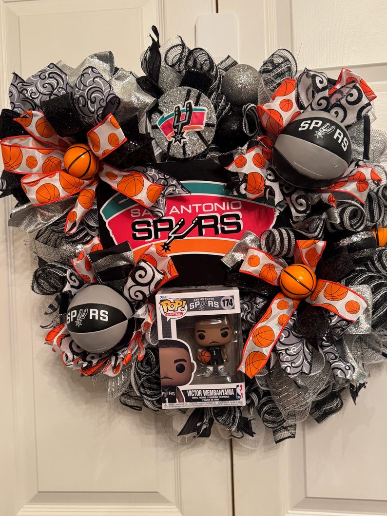 Puede incluir: Una corona decorativa con elementos del equipo de baloncesto de los San Antonio Spurs. La corona incluye cintas con tem&aacute;tica de baloncesto, adornos de baloncesto y una figura Funko Pop de Victor Wembanyama. La combinaci&oacute;n de colores incluye negro, plata, naranja y blanco.