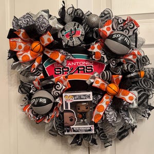 Puede incluir: Una corona decorativa con el tema de los Spurs de San Antonio. Incluye balones de baloncesto, cintas con los colores del equipo y una figura Pop! de Victor Wembanyama. La combinaci&oacute;n de colores incluye negro, plateado, naranja y blanco.