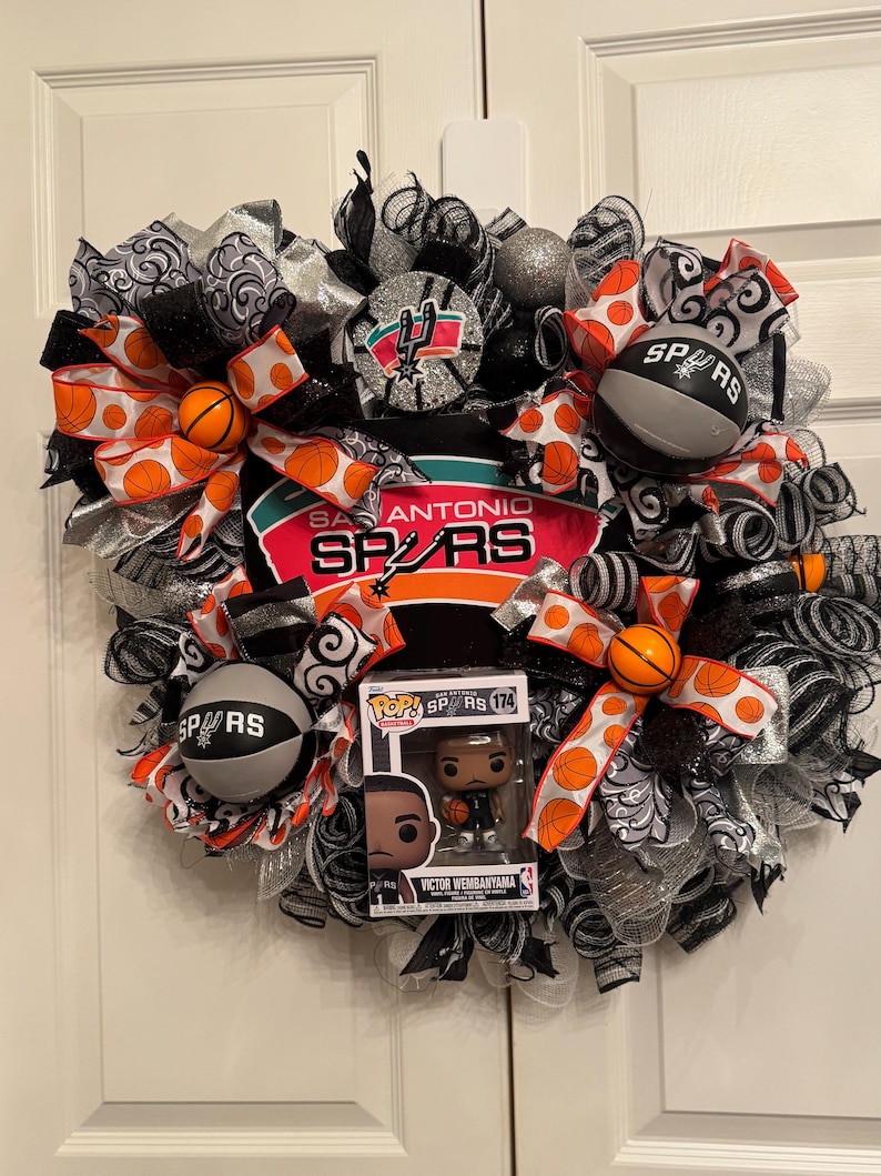 Puede incluir: Una corona decorativa con el tema de los Spurs de San Antonio. Incluye balones de baloncesto, cintas con los colores del equipo y una figura Pop! de Victor Wembanyama. La combinaci&oacute;n de colores incluye negro, plateado, naranja y blanco.