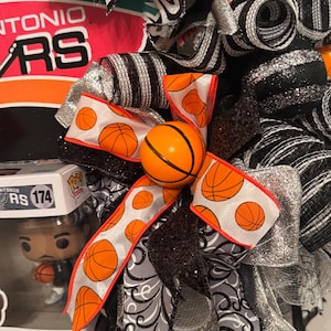 Puede incluir: Una corona decorativa con tem&aacute;tica de baloncesto, con cintas naranjas, negras, plateadas y blancas. La corona incluye un adorno de bal&oacute;n de baloncesto y el logotipo de los Spurs de San Antonio. Tambi&eacute;n hay una figura de Victor Wembanyama.