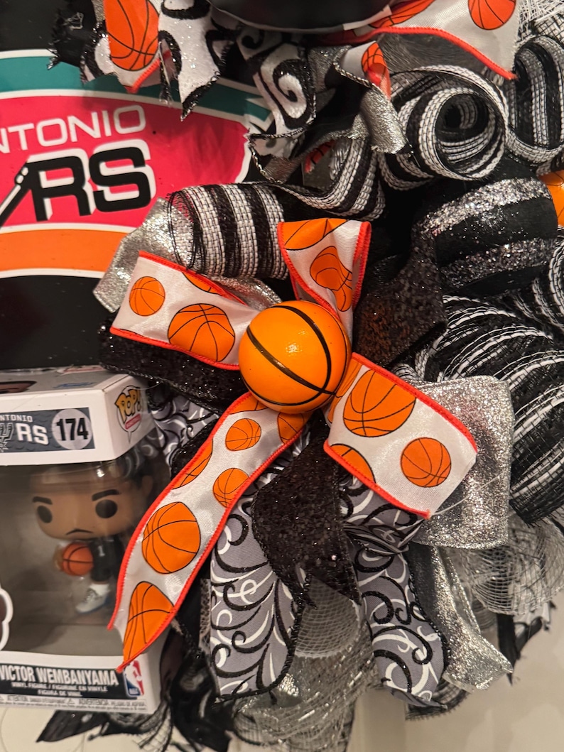 Puede incluir: Una corona decorativa con tem&aacute;tica de baloncesto, con cintas naranjas, negras, plateadas y blancas. La corona incluye un adorno de bal&oacute;n de baloncesto y el logotipo de los Spurs de San Antonio. Tambi&eacute;n hay una figura de Victor Wembanyama.