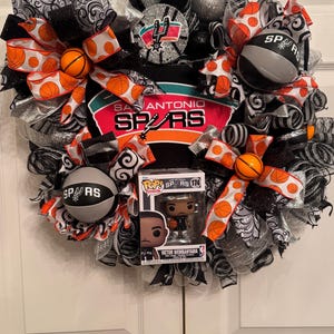 Puede incluir: Una corona decorativa con el logotipo y los colores de los San Antonio Spurs. La corona incluye detalles tem&aacute;ticos de baloncesto, cintas y una figura Funko Pop de Victor Wembanyama. La combinaci&oacute;n de colores incluye negro, plata, naranja y blanco.