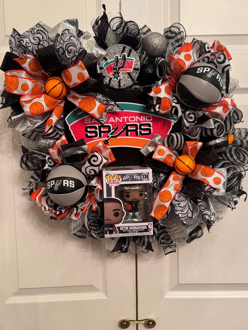 Puede incluir: Una corona decorativa con el logotipo y los colores de los San Antonio Spurs. La corona incluye detalles tem&aacute;ticos de baloncesto, cintas y una figura Funko Pop de Victor Wembanyama. La combinaci&oacute;n de colores incluye negro, plata, naranja y blanco.
