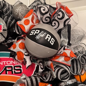 Puede incluir: Una corona decorativa con tem&aacute;tica de baloncesto. La corona incluye una pelota de baloncesto gris con la inscripci&oacute;n "SPURS", acentuada por cintas naranjas y negras con patrones de baloncesto y espirales. La malla plateada y negra completa el dise&ntilde;o.