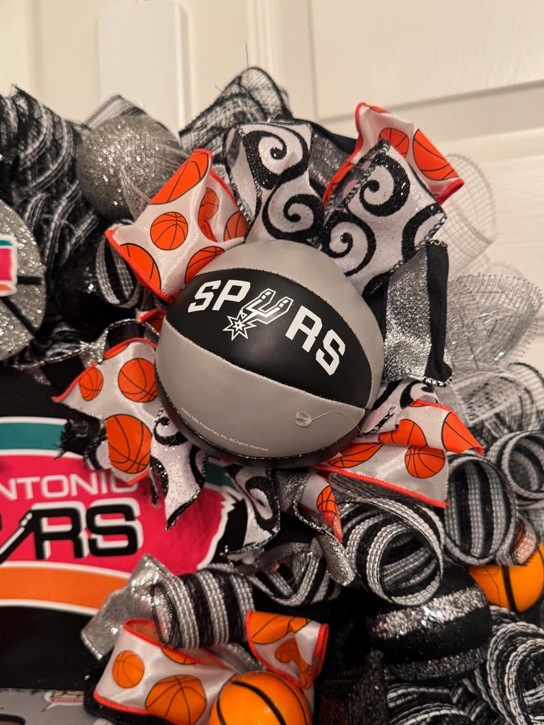 Puede incluir: Una corona decorativa con tem&aacute;tica de baloncesto. La corona incluye una pelota de baloncesto gris con la inscripci&oacute;n "SPURS", acentuada por cintas naranjas y negras con patrones de baloncesto y espirales. La malla plateada y negra completa el dise&ntilde;o.