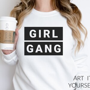 Può includere: Felpa bianca con testo nero che recita "GIRL GANG" in un carattere audace e a blocchi. Il testo è centrato sulla felpa. La felpa è indossata da una persona che tiene una tazza di caffè.