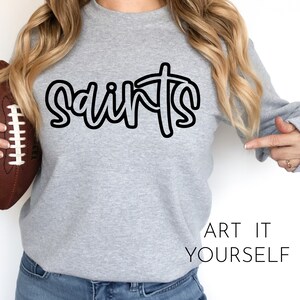 以下が含まれることがあります： グレーのトレーナーに、黒い筆記体で「Saints」と書かれています。「Saints」という単語は、単語の真ん中に十字架が描かれたスタイリッシュなデザインです。画像の下部には、「Art It Yourself」というテキストが黒で印刷されています。
