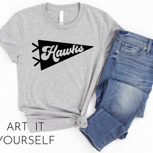 May include: A gray t-shirt with a black pennant design that says "Hawks" on it. The t-shirt is paired with blue jeans.