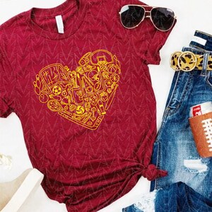 May include: Burgundy t-shirt with a yellow heart-shaped design composed of various sports equipment. The shirt is paired with blue jeans, a leopard print belt, sunglasses, and a football-shaped can cooler.