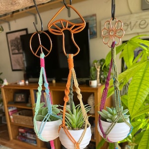 May include: Three macrame plant hangers with white ceramic pots, each holding a succulent. The hangers are suspended from a branch, featuring copper wire designs of a peace sign, mushroom, and flower. The macrame cords are in various colors.