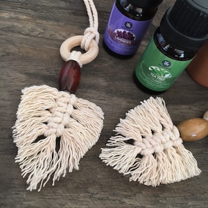May include: Macrame car air freshener with a wooden ring and bead, paired with essential oil bottles labeled "Lavender" and "Tea Tree". The macrame design features a leaf shape in a natural, off-white color.