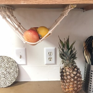 May include: A white macrame hammock hanging from a wooden shelf, holding a red apple, a yellow lemon, and a pineapple on a kitchen counter.