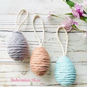 Peut inclure: Trois &oelig;ufs de P&acirc;ques faits main, chacun envelopp&eacute; dans une couleur de fil diff&eacute;rente. Les &oelig;ufs sont gris, p&ecirc;che et bleu clair. Les &oelig;ufs sont suspendus &agrave; une ficelle blanche.