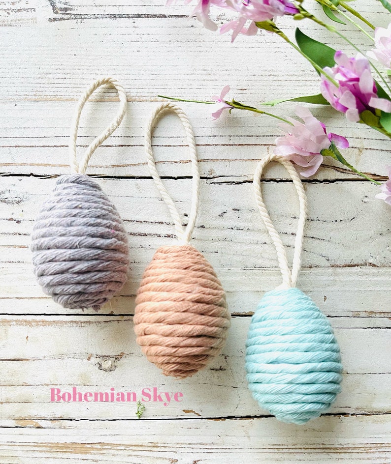 Peut inclure: Trois &oelig;ufs de P&acirc;ques faits main, chacun envelopp&eacute; dans une couleur de fil diff&eacute;rente. Les &oelig;ufs sont gris, p&ecirc;che et bleu clair. Les &oelig;ufs sont suspendus &agrave; une ficelle blanche.