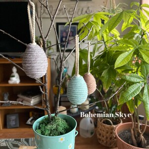 Peut inclure: Trois &oelig;ufs d&eacute;coratifs en corde, suspendus &agrave; une branche nue dans un pot vert avec de la mousse. Les &oelig;ufs sont gris, bleu clair et marron. Le pot a un motif de marguerite blanche. Le texte "Bohemian Skye" est visible sur l'image.
