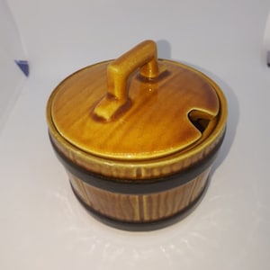May include: A ceramic jar with a lid and handle. The jar is a warm brown color with a dark brown band around the middle. The lid has a handle and a small notch. The jar has a rustic, barrel-like design.