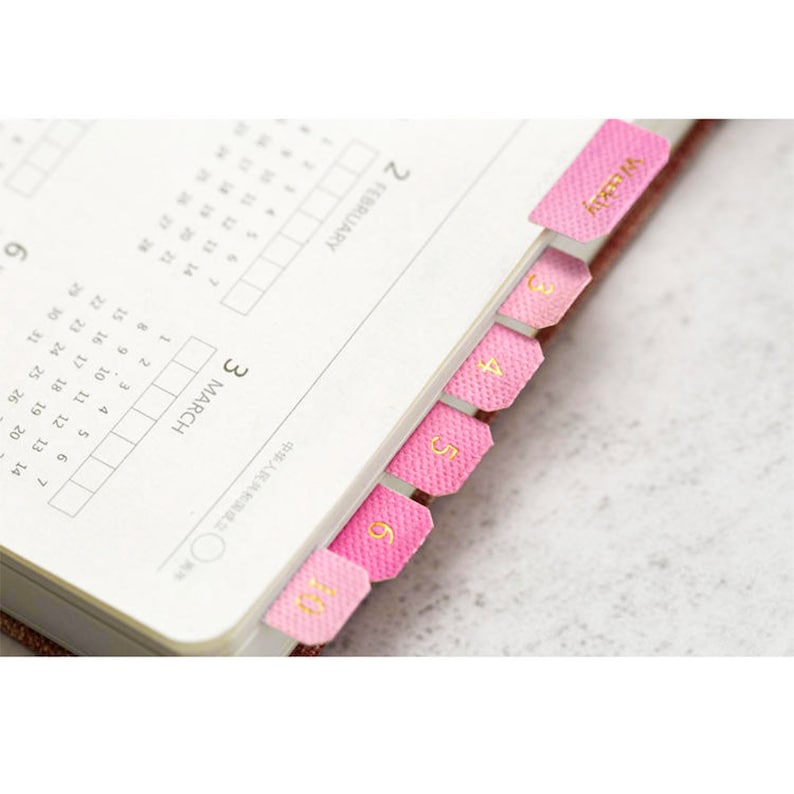 May include: Pink page markers with gold numbers 1 through 7 for a planner or calendar. The markers are labeled "Weekly" and "February".