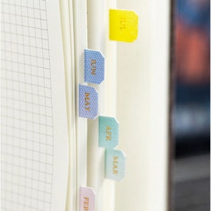May include: A set of six colorful page markers with month names in gold lettering. The markers are in shades of pink, green, blue, and yellow. The months are February, March, April, May, June, and July.