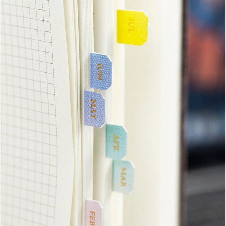 May include: A set of six colorful page markers with month names in gold lettering. The markers are in shades of pink, green, blue, and yellow. The months are February, March, April, May, June, and July.