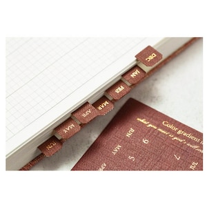 May include: A brown leather-bound planner with gold-colored tabs for each month of the year. The tabs are labeled January through December.