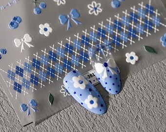 May include: A sheet of adhesive nail art stickers featuring blue and white patterns including argyle strips, small flowers, bows, and cherries. Two light blue almond-shaped nail tips are displayed, one with white flower accents and one with a white bow.