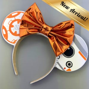 May include: A headband with white ears featuring orange and white graphics, a large metallic orange bow, and the text "New Arrival!". The headband is white with a pearl bead accent.