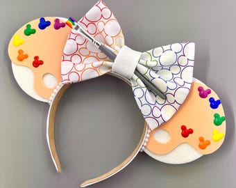 May include: A pair of white mouse ears with a rainbow Mickey Mouse print bow and a paint palette with colourful paint drops. The ears are attached to a brown headband.