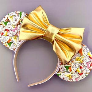 May include: A pair of mouse ears with a gold bow and a white headband. The ears are decorated with a colorful pattern of cartoon characters. The bow is made of shiny gold material.