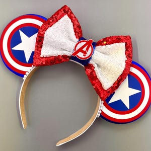 Puede incluir: Una diadema con temática de Capitán América. Cuenta con dos escudos circulares con una estrella blanca sobre fondo azul, detalles en rojo y blanco, y un lazo de lentejuelas con el logotipo de los Vengadores. La diadema es blanca con pequeñas perlas.