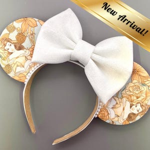 Puede incluir: Una diadema con lazo blanco y orejas de ratón. Las orejas presentan un diseño de Bella con detalles florales. La diadema tiene un ribete de perlas y el texto "New Arrival!" es visible.