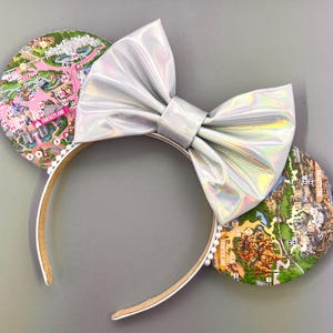 Puede incluir: Un par de orejas de ratón blancas con un lazo plateado iridiscente. Las orejas están decoradas con un mapa de un parque temático, incluyendo el texto "Fantasyland".