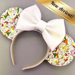 May include: A white headband with attached mouse ears featuring a colorful pattern of cartoon characters. A large white bow sits atop the headband. The text "New Arrival!" is displayed on a gold banner.