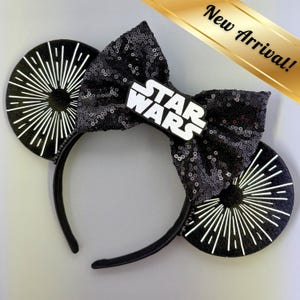 May include: Black sequined headband with a bow and two round ears. The bow features a white "Star Wars" logo. The ears have a white burst design. A gold banner in the top right corner reads "New Arrival!"