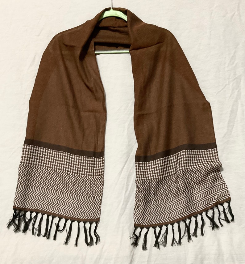 May include: A brown scarf with a white and brown geometric pattern on the ends. The scarf has black tassels.